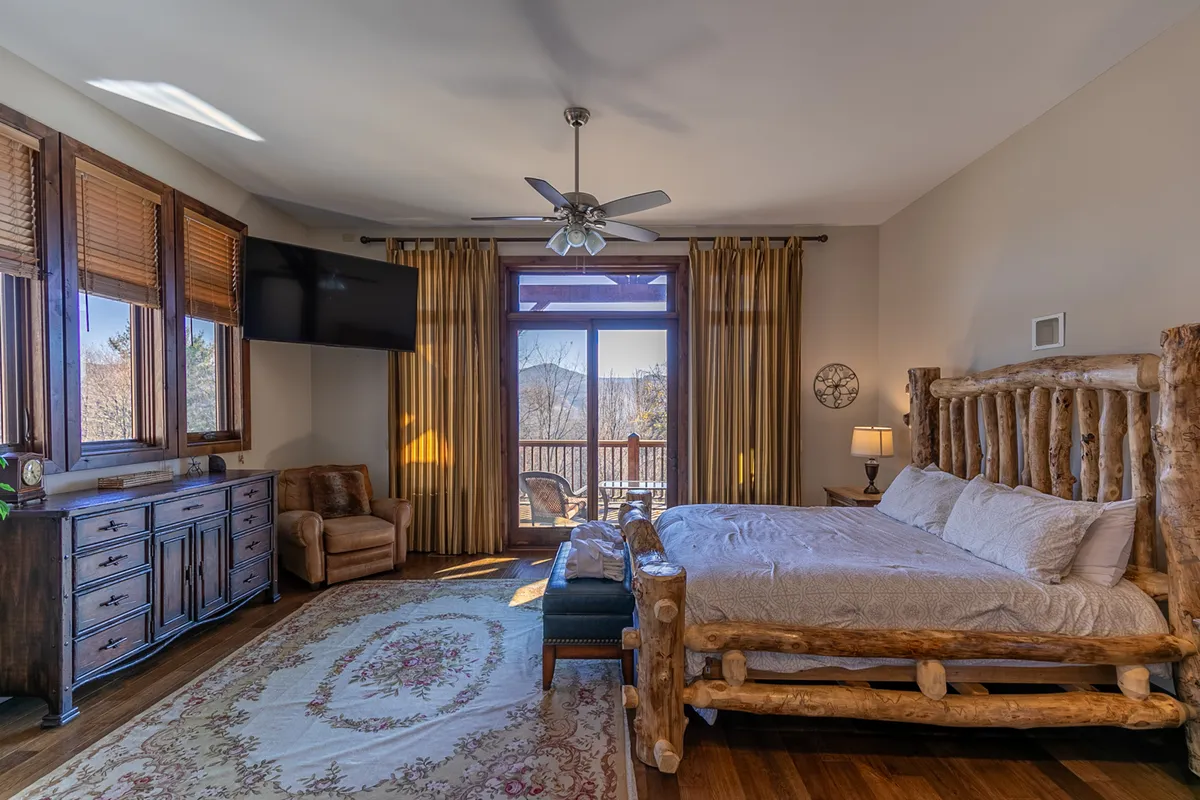 This image depicts a serene bedroom with a log bed, a large window offering mountain views, a wooden dresser, and a comfortable chair set atop an area rug. The room is bathed in natural light, conveying a sense of calm and tranquility ideal for restful retreats.