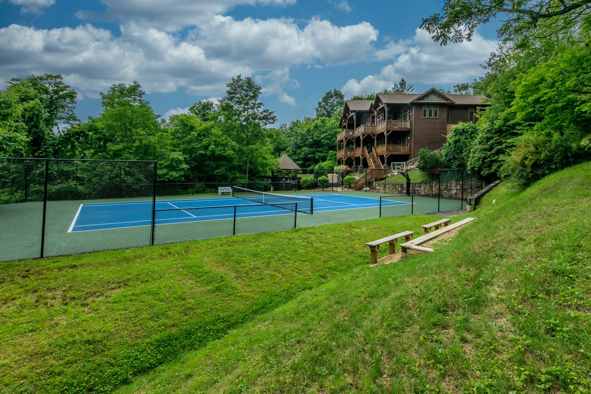The photo showcases a private hardtop tennis court bordered by lush greenery, with a multi-story lodge in view. Benches for spectators and a spacious lawn suggest space for friendly matches and outdoor gatherings, all within a serene estate setting.