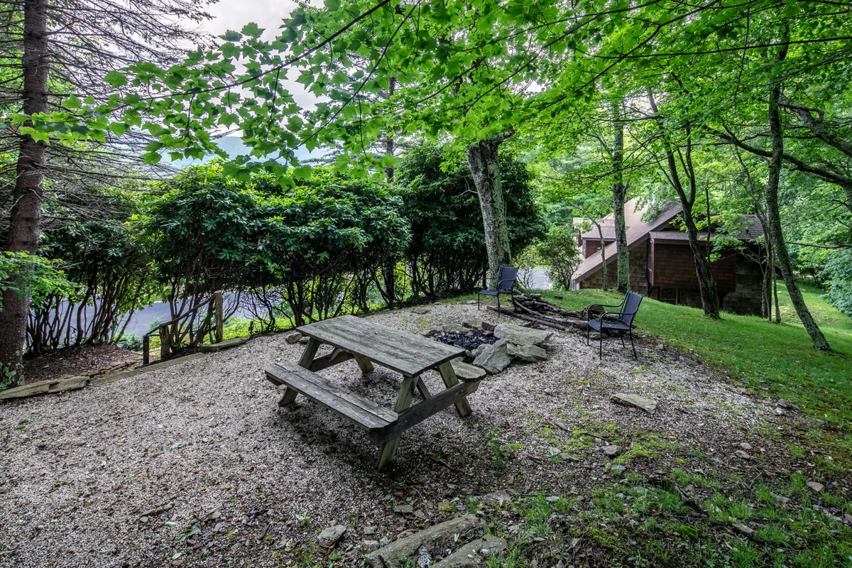 A picturesque outdoor scene with a picnic table and a stone fire pit encircled by lawn chairs, set amidst lush greenery. The area invites family gatherings, storytelling, and peaceful evenings under the stars.