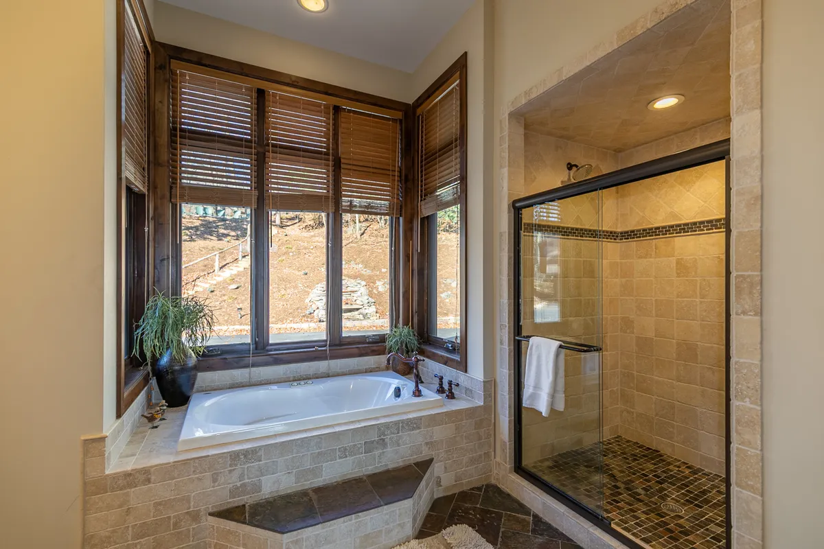A luxurious bathroom dominated by a spacious bathtub set against large windows with natural light streaming in, complemented by a roomy shower area. The setting offers a serene, spa-like ambiance for unwinding.