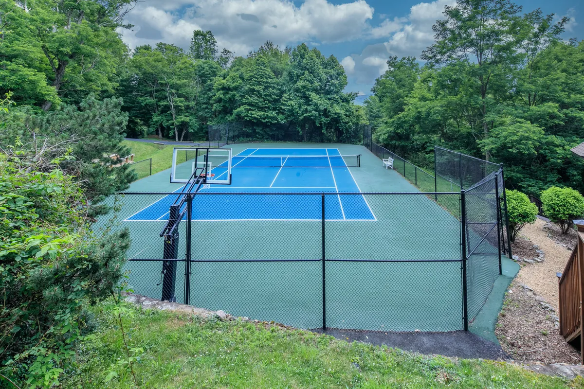 A vibrant exterior view featuring the tennis court complete with a basketball hoop, framed by rich greenery. The area is bathed in sunlight, suggesting active days spent outdoors playing both tennis and basketball.