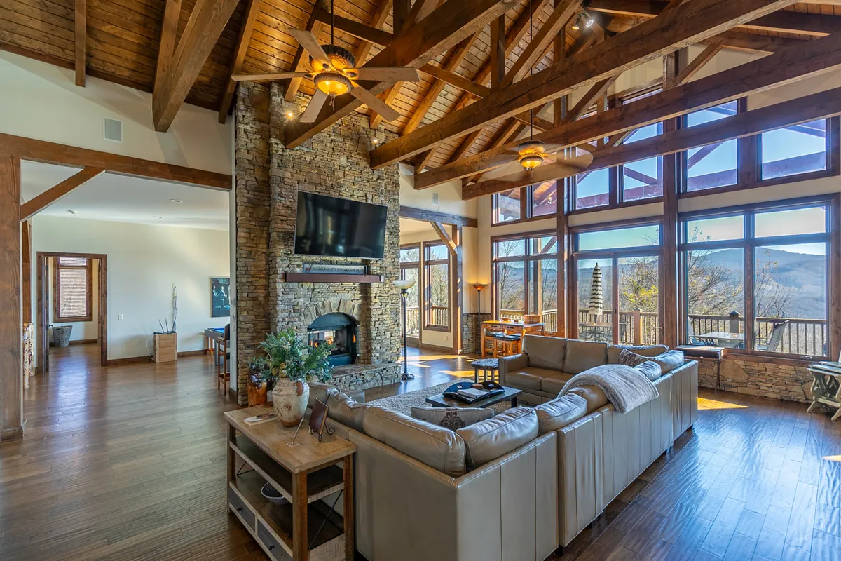A grand great room featuring large panoramic windows that flood the space with natural light, a striking stone fireplace, exposed wooden beams, and plush comfortable seating arranged for gatherings. The setting is cozy yet elegant, ideal for both lively celebrations and quiet, relaxing evenings.