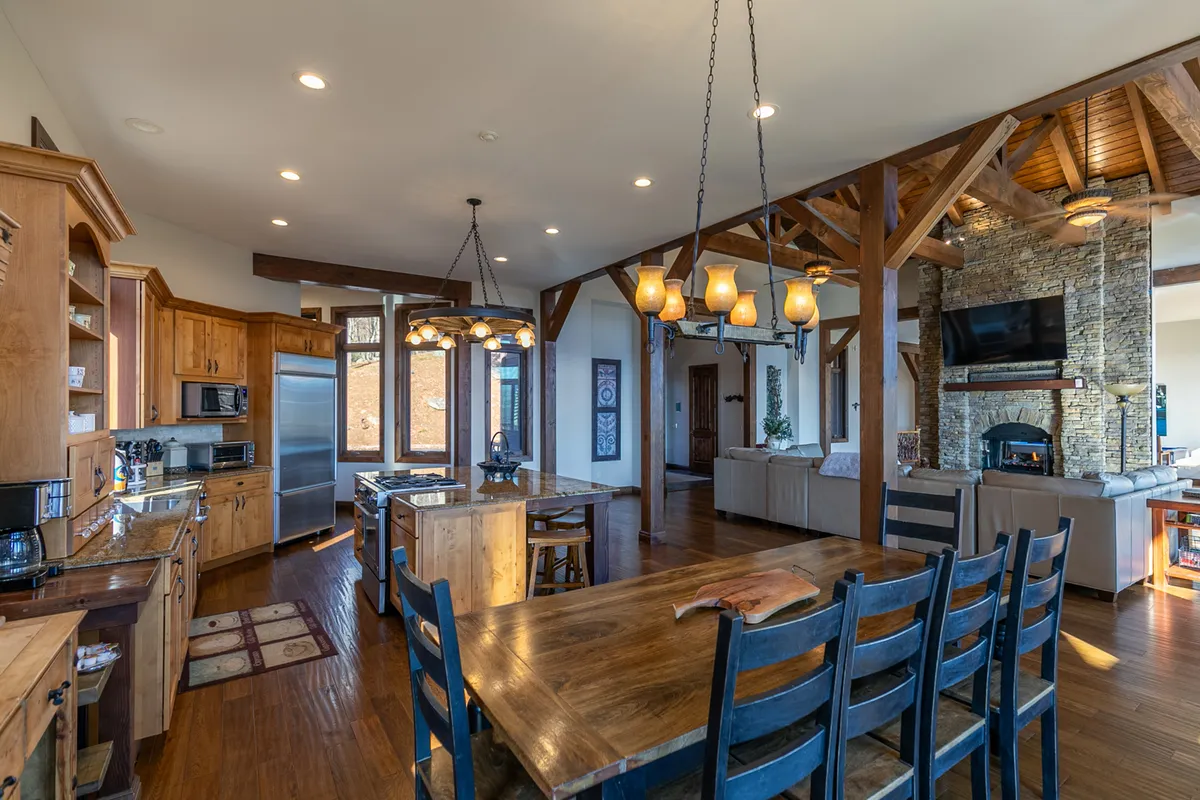 A warm, inviting dining space features a large wooden dining table, an island kitchen with bar stools, exposed wooden beams, a stone fireplace, and expansive windows offering scenic views. The setup is ideal for family gatherings and memorable dinner parties.