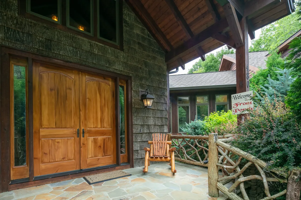 A close-up exterior shot of rustic wooden double doors, a stone pathway, a welcoming rocking chair, and decorative lanterns on the porch. The scene feels warm and inviting, ideal for arrivals and cozy evenings.