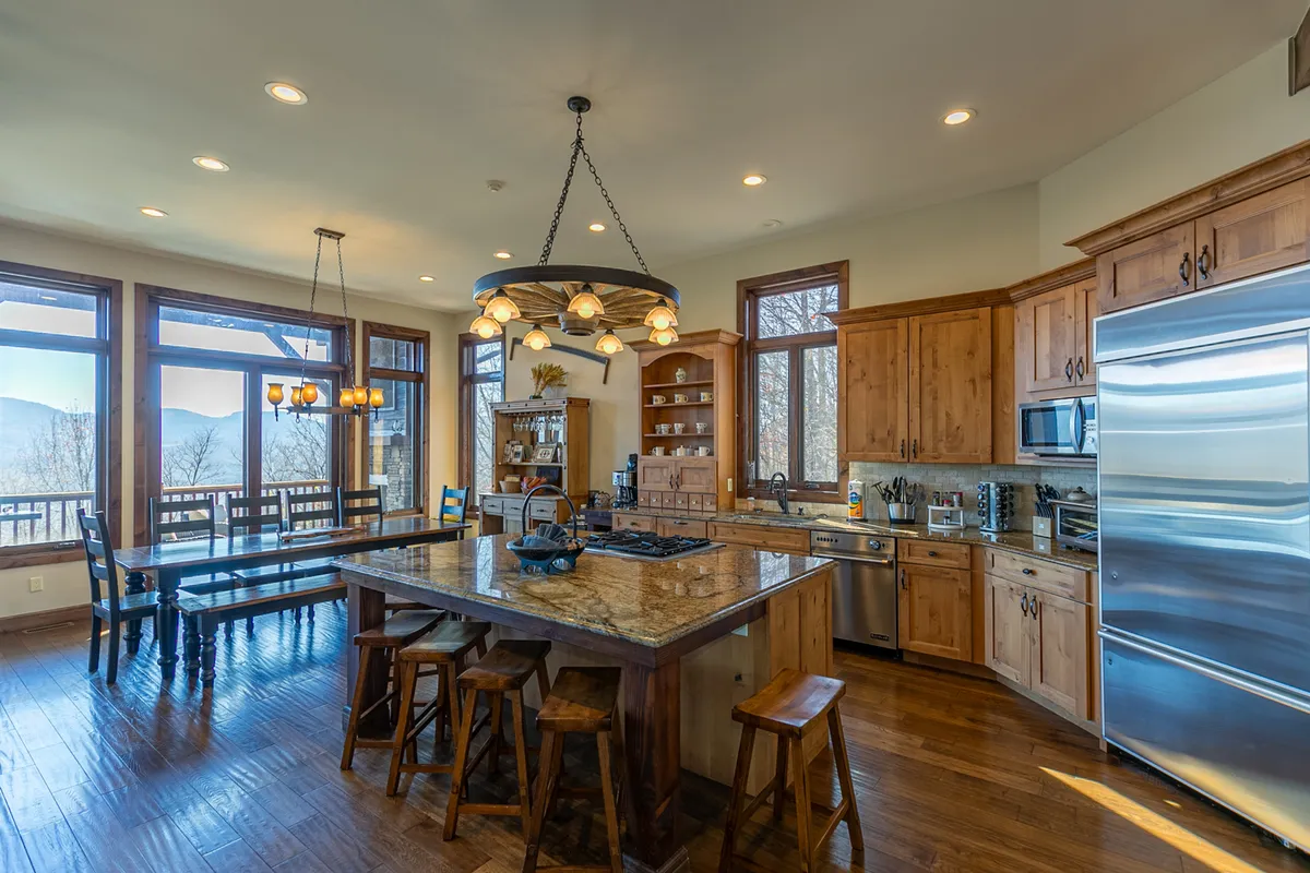 A bright, spacious kitchen featuring a large kitchen island, wooden cabinets, panoramic windows, and a nearby dining table beneath an overhead chandelier. The open layout is filled with natural light and showcases both the kitchen and dining spaces, with plenty of room for gathering and meal preparation.