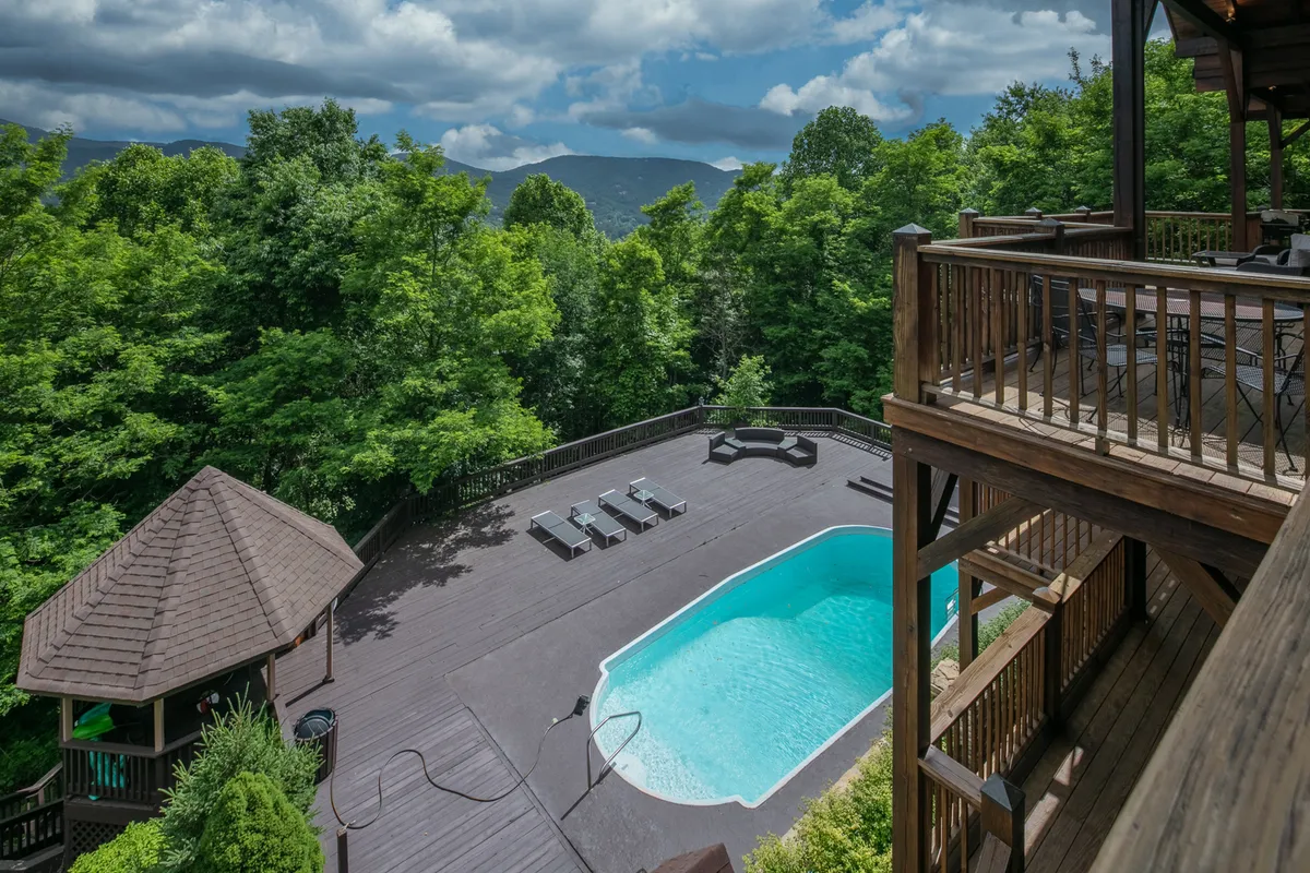 A tranquil pool area with a large deck, surrounded by trees and mountain views. The scene includes a shaded wooden gazebo near the pool, offering a peaceful spot for relaxing or socializing, and ample outdoor space for entertaining.